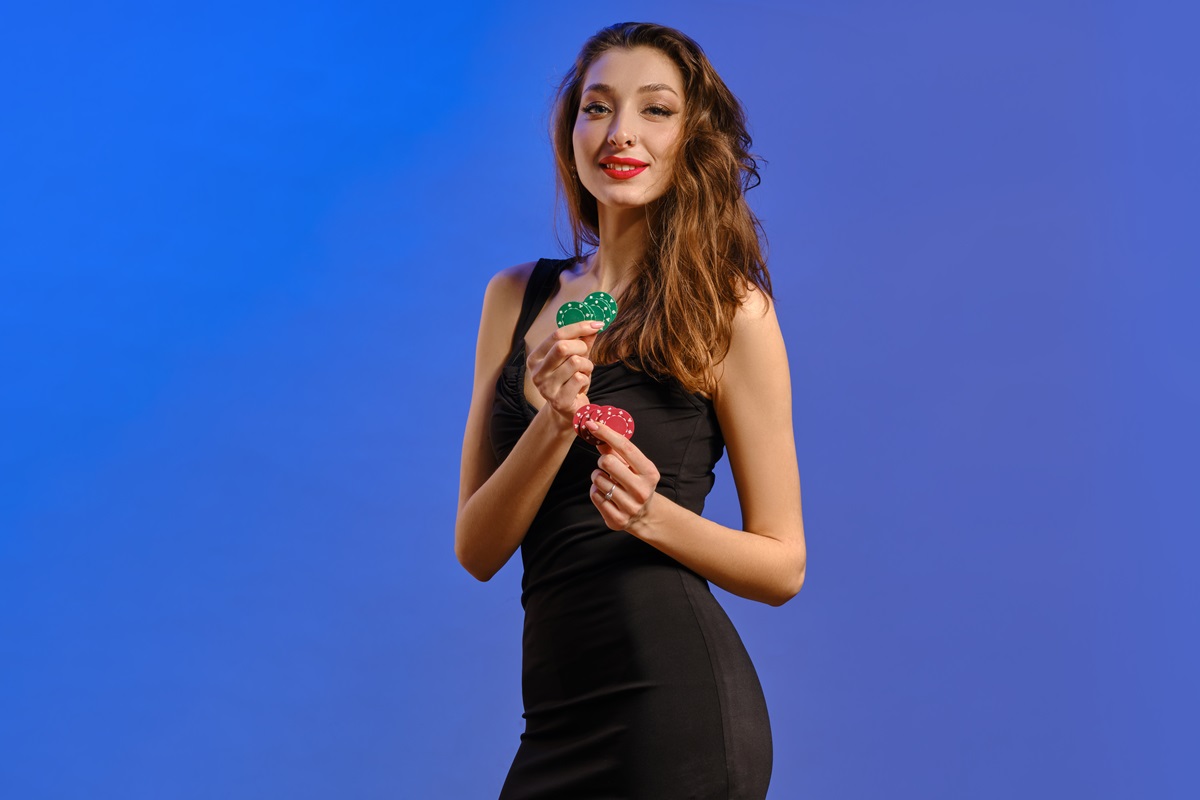 Brunette model with earring in nose, in black dress. She smiling, holding two green and red chips, posing on blue background. Poker, casino. Close-up