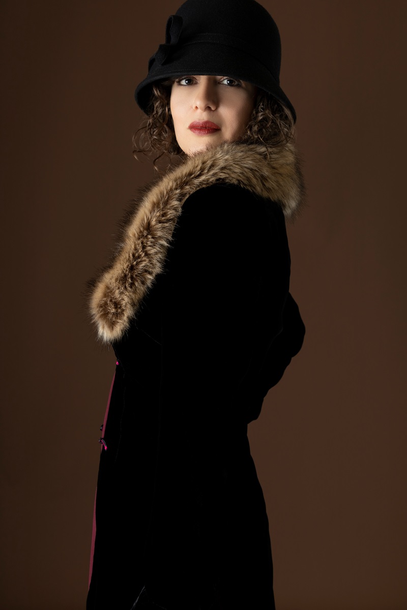 A 1920s woman wearing a black velvet coat with a fur collar and standing against a plain studio backdrop